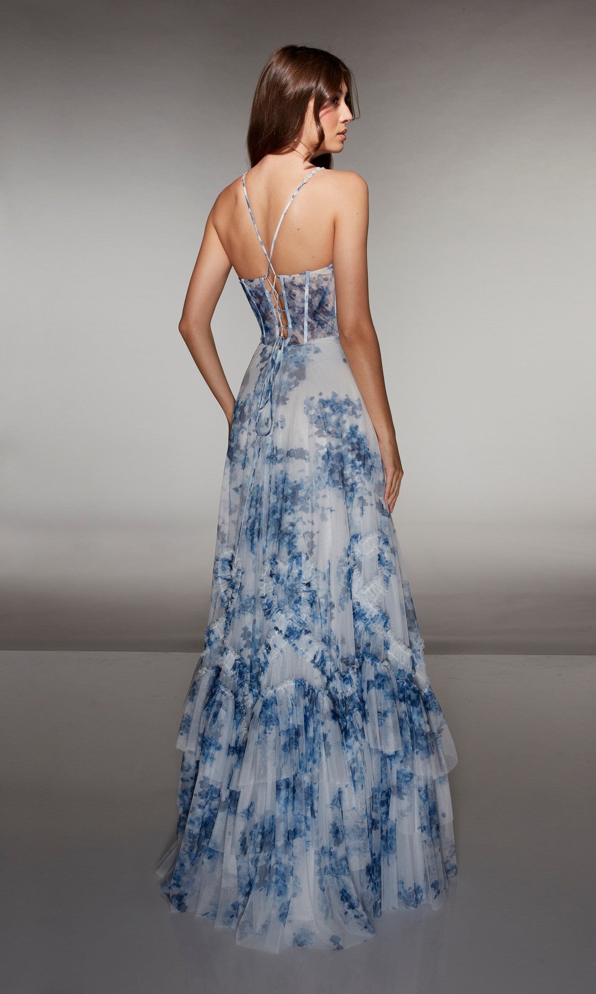A woman faces away, wearing the Alyce 62028 long prom dress featuring blue-and-white floral print, thin straps, a sheer corset bodice, and tiered layers against a neutral background.